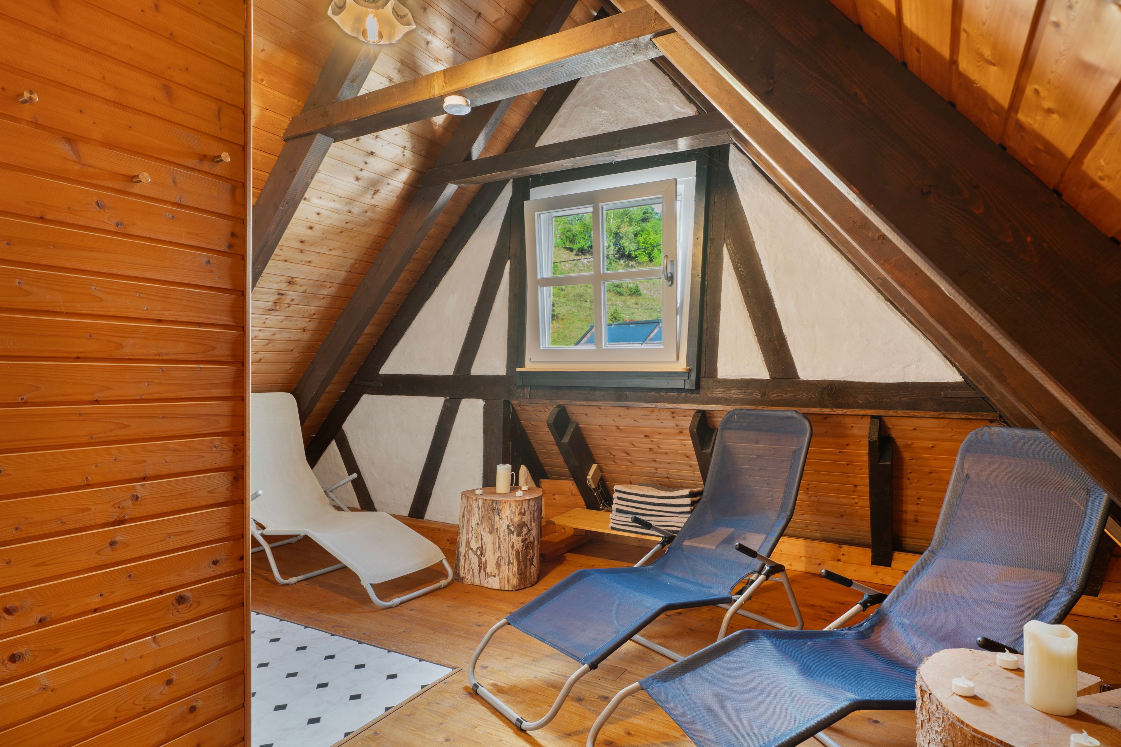 A cozy attic room with wooden beams, a window with a view, and two blue chairs.
