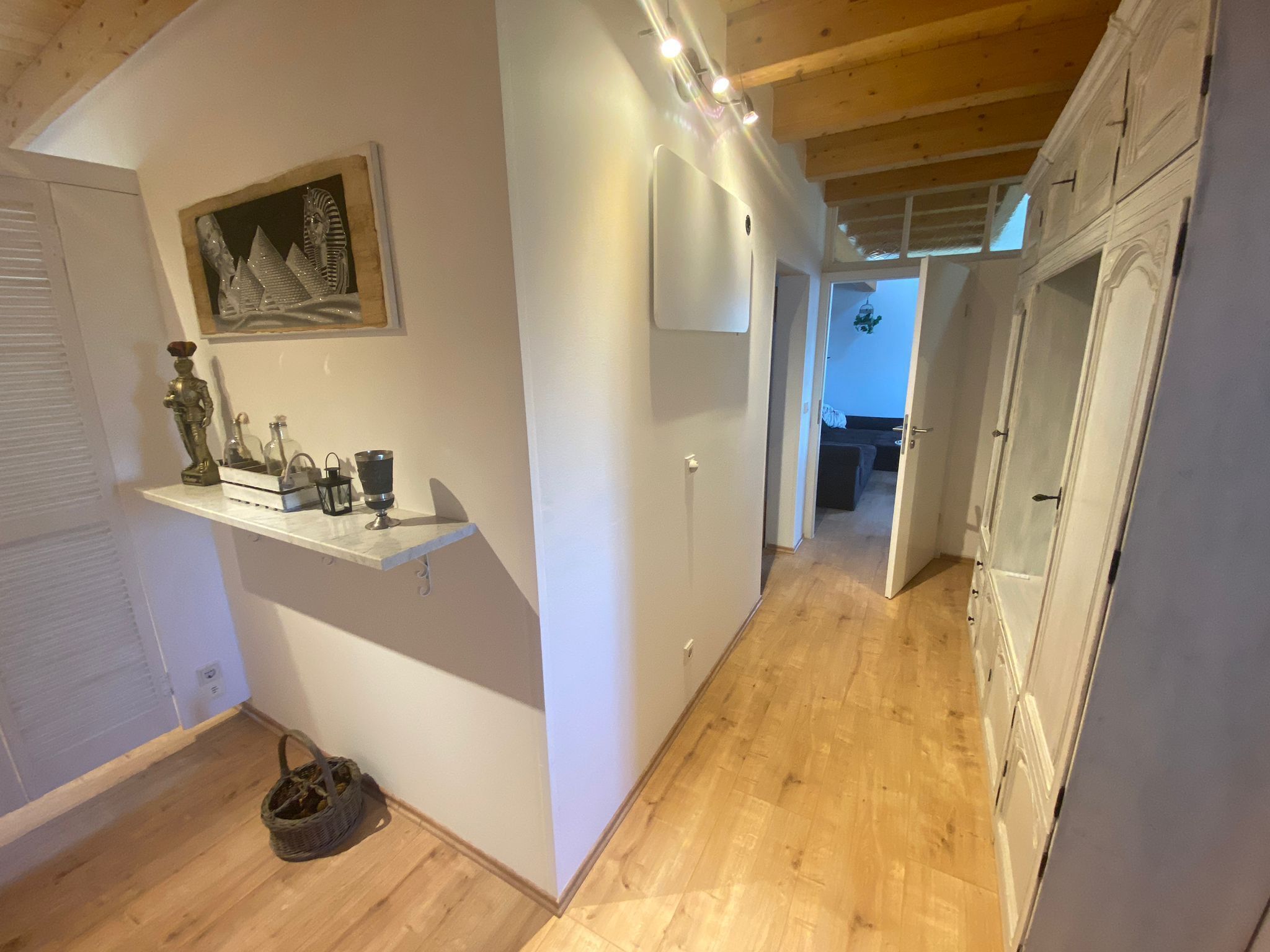 Un couloir doté d’un plancher en bois, de murs blancs, d’une étagère ornée d’objets décoratifs et d’une porte donnant sur une autre pièce.