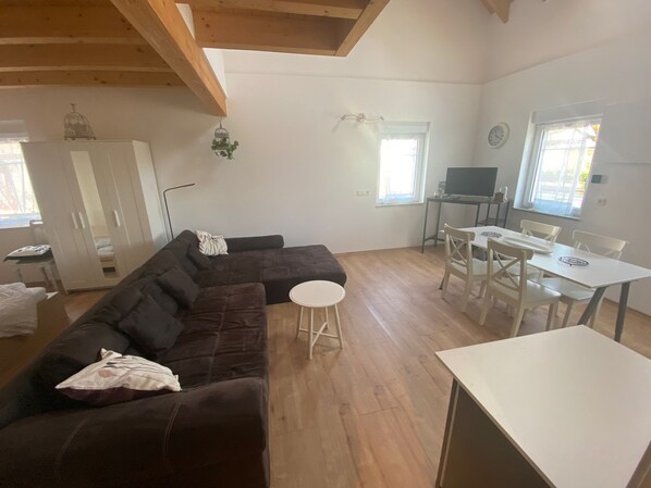 A living room with a dark brown sectional sofa, a white dining table with four chairs, a small white stool, and a TV on a stand. - Idyll of the Vulkaneifel, vacation apartment in Laufeld (Laufeld)