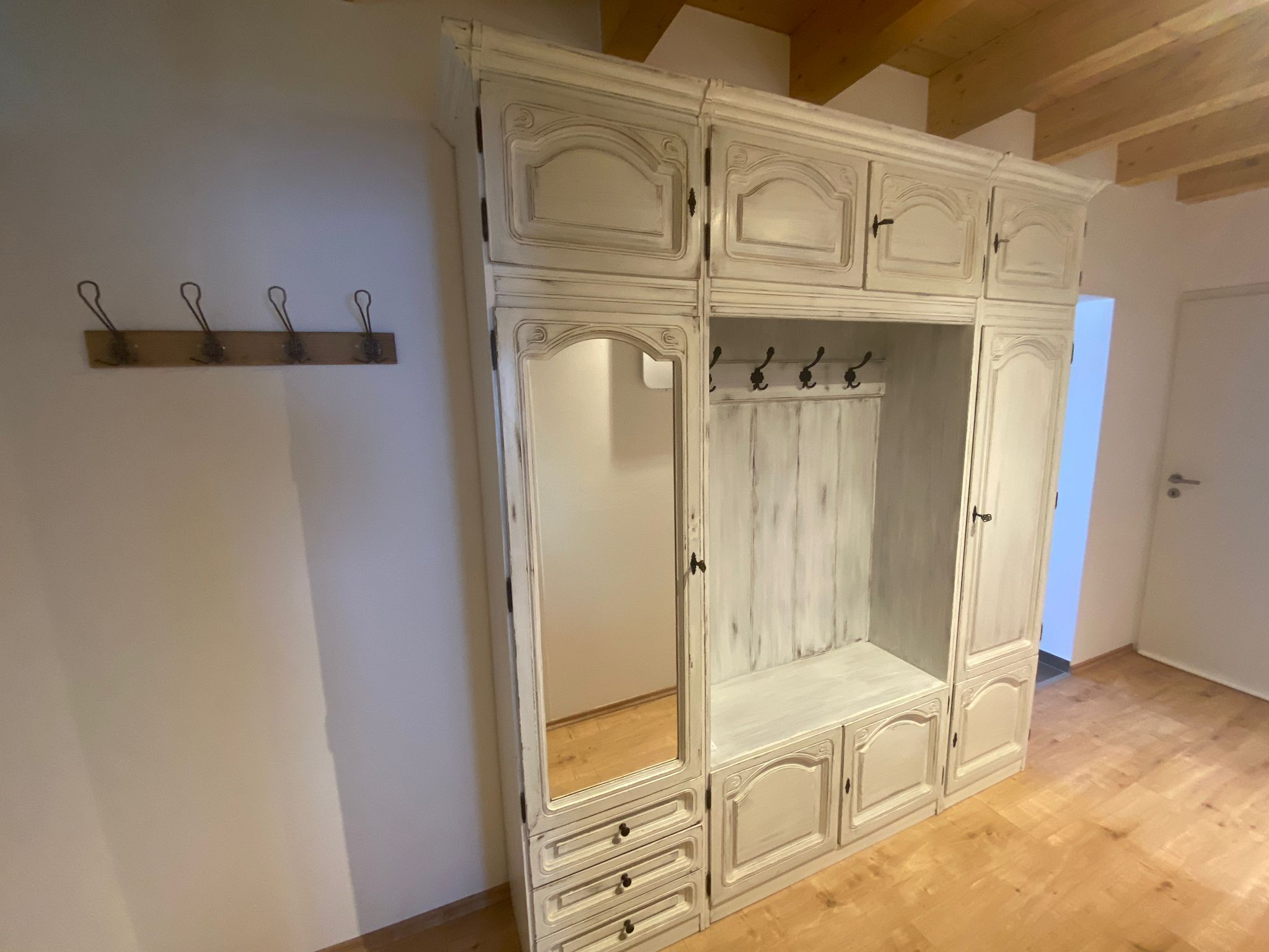 Ein großer, weißer Kleiderschrank mit mehreren Türen und Schubladen, ein Holzregal mit Haken sowie ein Holzboden.