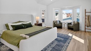 A bedroom with a large bed, a sofa, a chair, a small table, and a view of buildings through a window. - Outrigger Beach Retreat w/ Private Path & Firepit (Oxnard)