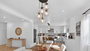 A modern kitchen with white cabinets, a wooden dining table set with plates and candles, and a hanging light fixture. - Outrigger Beach Retreat w/ Private Path & Firepit (Oxnard)