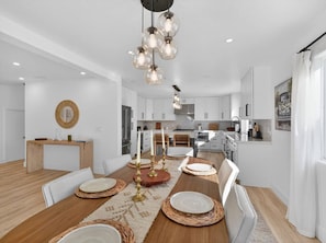 A modern kitchen with white cabinets, a wooden dining table set with plates and candles, and a hanging light fixture. - Outrigger Beach Retreat w/ Private Path & Firepit (Oxnard)
