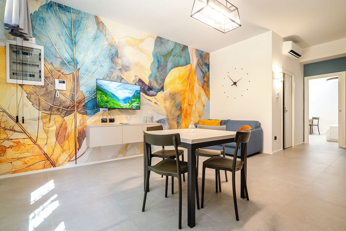 A modern living room with a dining area, a flat-screen TV, and a large abstract leaf mural on the wall.