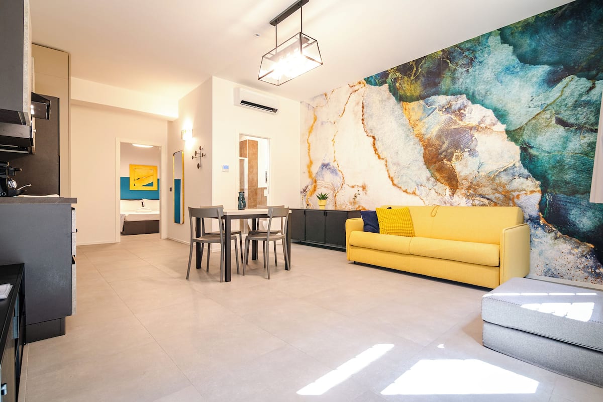 A modern living room with a yellow sofa, a dining table with chairs, and a large abstract painting on the wall.