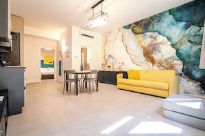 A modern living room with a yellow sofa, a dining table with chairs, and a large abstract painting on the wall.