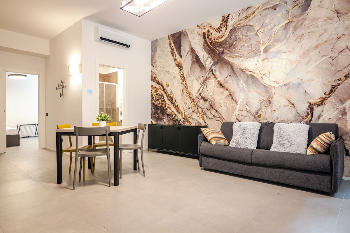 A living room with a sofa, a dining table, chairs, and a large marble-patterned wall.
