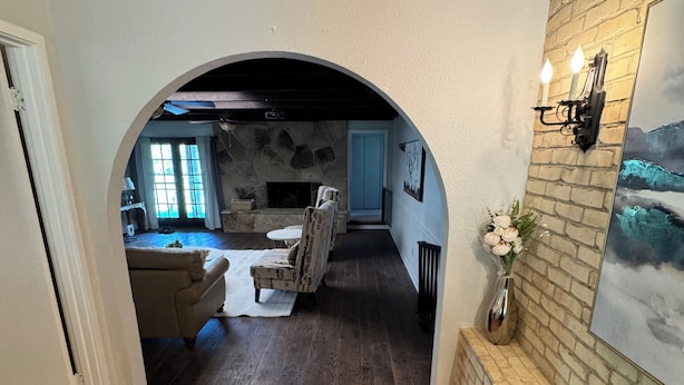 An arched doorway leading to a living room with a fireplace, a dining area with a table and chairs, and a brick wall with a painting.