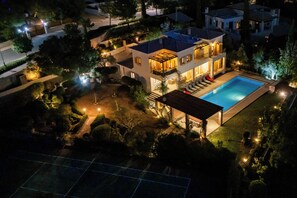 Ein modernes, zweistöckiges Haus mit Swimmingpool, umgeben von angelegten Gärten und einem Tennisplatz.