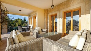 A covered outdoor seating area with wicker furniture and a glass table, stone walls, and a view of trees. - Villa 151 Luxury Villa - Private Pool and Tennis Court (Kouklia)