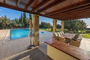 A covered outdoor area with a pool, stone pillars, a dining table with chairs, and a view of the surrounding landscape. - Villa 151 Luxury Villa - Private Pool and Tennis Court (Kouklia)