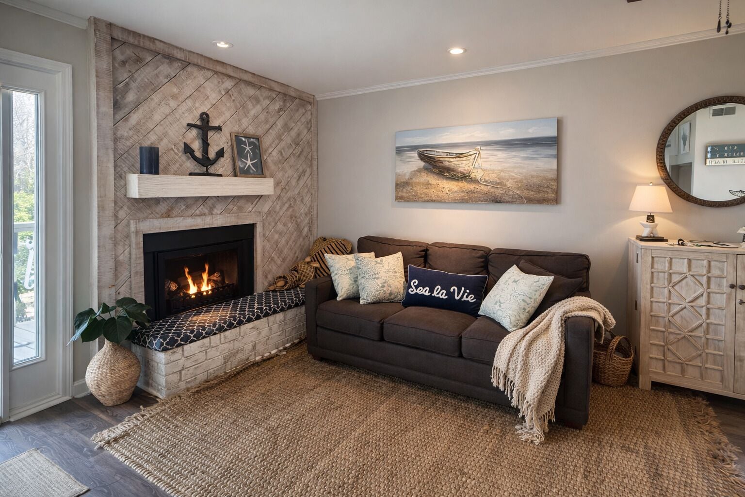 Una sala de estar con chimenea, un sofá con cojines decorativos, un cuadro de un barco en el agua y un espejo redondo sobre un aparador.