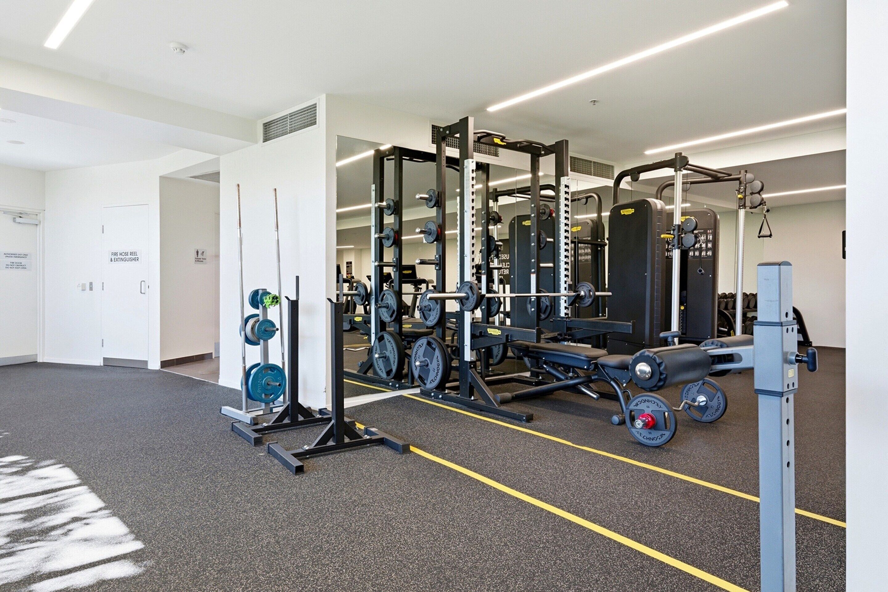 A gym with weightlifting equipment, a door labeled 'Fire Hose Reel & Extinguisher,' and a carpeted floor.