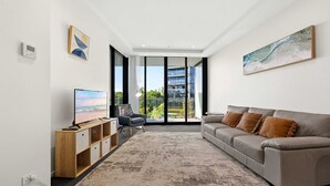 En moderne stue med sofa, fjernsyn, stol og et stort vindue med udsigt til en bygning og grønne områder.