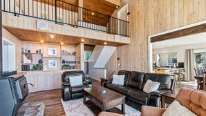 A spacious living area with a high ceiling, wooden walls, a fireplace, leather sofas, and a kitchen in the background.