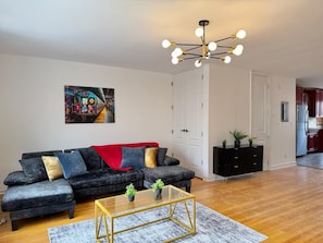 Ein Wohnzimmer mit Ecksofa, Glastisch, moderner Pendelleuchte, einem Gemälde an der Wand und einem durchgehenden Türrahmen erkennbaren Küchenbereich.