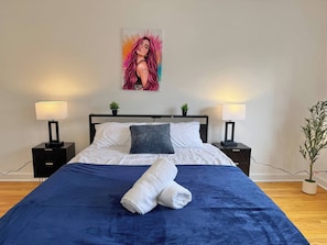 Ein ordentlich bezogenes Bett mit blauer Bettdecke und weißen Kissen, zwei Nachttische mit Lampen, eine kleine Pflanze auf jedem Tisch und ein Bild an der Wand.