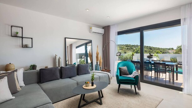 Uma sala de estar moderna com um sofá cinzento, uma mesa de centro redonda, uma poltrona verde-água e uma grande janela com vista para uma piscina e vegetação.