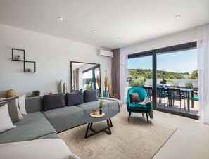 A modern living room with a grey sofa, a round coffee table, a teal armchair, and a large window offering a view of a pool and greenery. - Stay in Kanica With Sauna (Rogoznica)