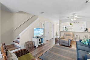 Una sala de estar con televisor, sofá, sillón, escalera y una zona de cocina.