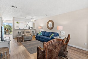 Sala de estar con sofá, sillones, una silla de mimbre, televisor, zona de cocina y balcón.