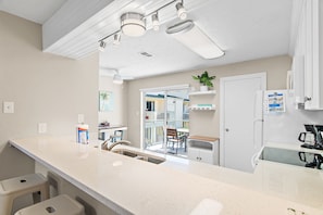 A kitchen with a white countertop, stainless steel sink, and a refrigerator. There is a window with a view of a house and a door leading to a balcony.