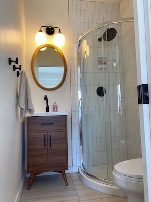 A bathroom with a wooden vanity, a round mirror, a glass shower enclosure, and a toilet.