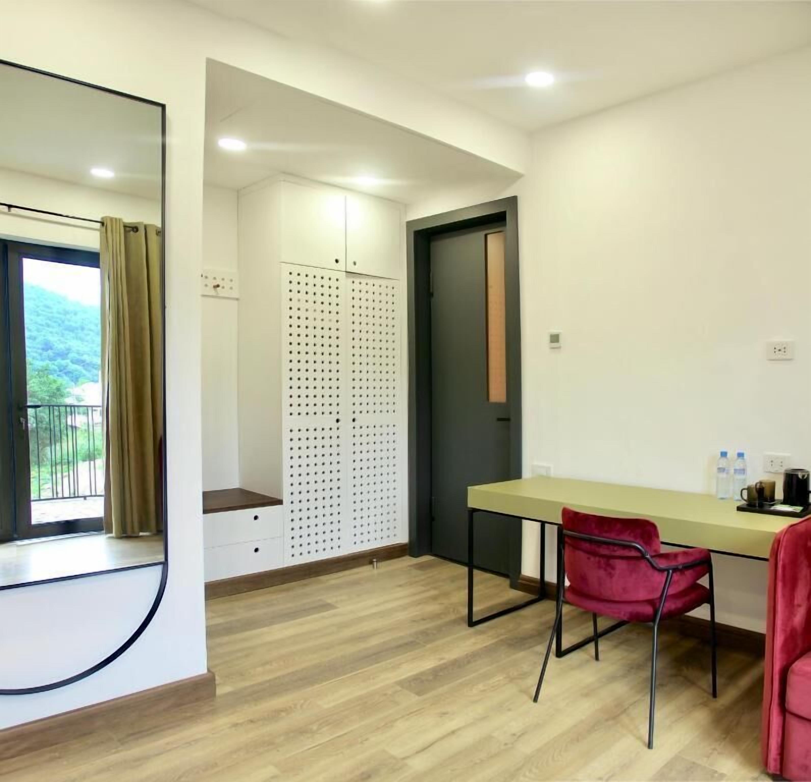 Um quarto com escrivaninha, cadeira, janela com cortinas, porta e espelho fixado na parede.