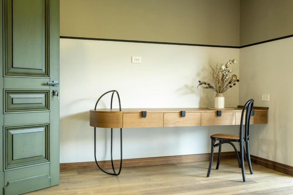 A room with a desk, chair, and a vase with dried plants on the wall.