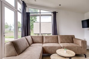 A modern living room with a sectional sofa, a round wooden coffee table, and a large window with curtains. - Lessenaar 22 | 6 persoons chalet in Zennewijnen (Zennewijnen)