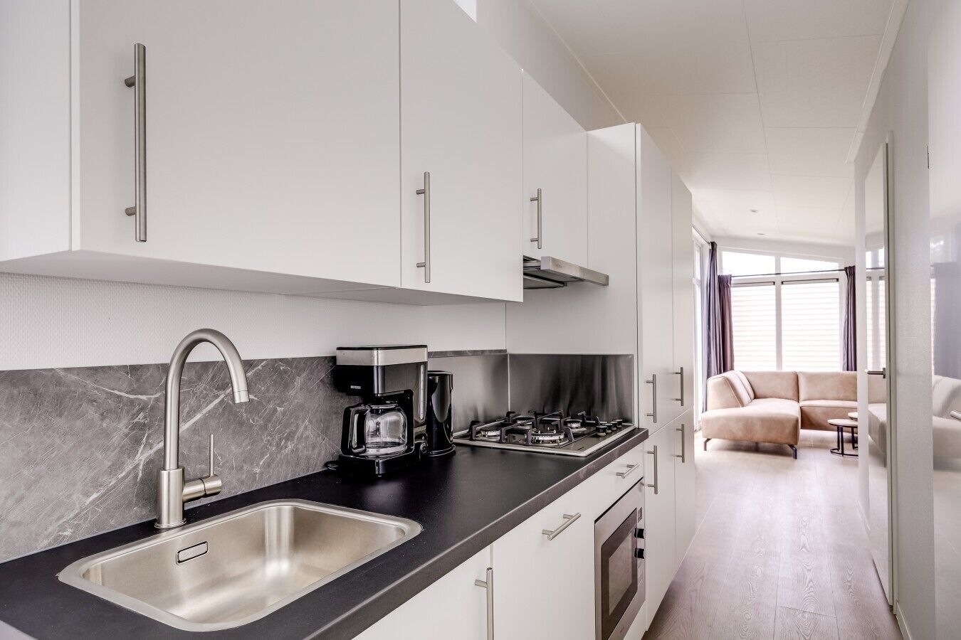 A modern kitchen with a stainless steel sink, black countertop, and white cabinets. There is a gas stove, a coffee maker, and a microwave. The kitchen leads to a living area with a beige sofa and a small table.