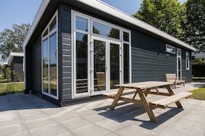 A modern house with a large sliding glass door, a wooden deck, and a bench. - Lessenaar 22 | 6 persoons chalet in Zennewijnen (Zennewijnen)