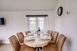 A dining area with a round table set for six, six upholstered chairs, a wall-mounted clock, and a window with blinds. - Lessenaar 22 | 6 persoons chalet in Zennewijnen (Zennewijnen)