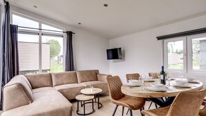 A modern living room with a sectional sofa, a round dining table set for four, and a flat-screen TV mounted on the wall. - Lessenaar 22 | 6 persoons chalet in Zennewijnen (Zennewijnen)