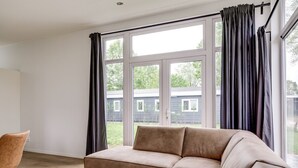 A modern living room with a beige sofa, a round coffee table, and a view of a house through large windows. - Lessenaar 22 | 6 persoons chalet in Zennewijnen (Zennewijnen)