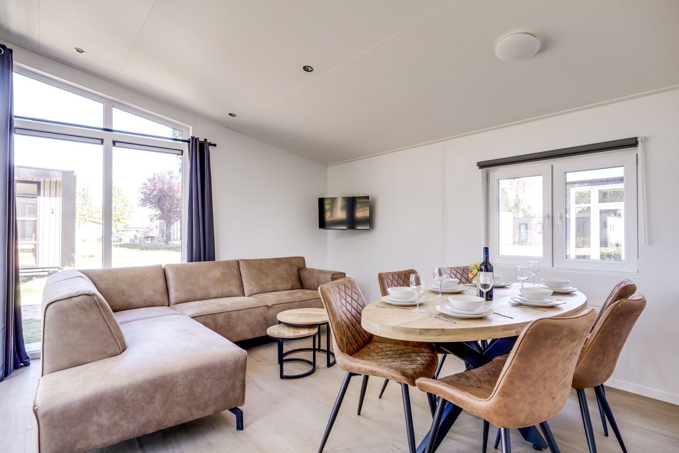 Ein modernes Wohnzimmer mit einem beigefarbenen Ecksofa, einem runden Esstisch mit Stühlen und großen Fenstern, die für natürliches Licht sorgen.