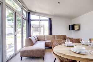 A modern living room with a sectional sofa, a dining table set for two, and a large window providing natural light. - Lessenaar 23 | 6 persoons chalet in Zennewijnen (Zennewijnen)