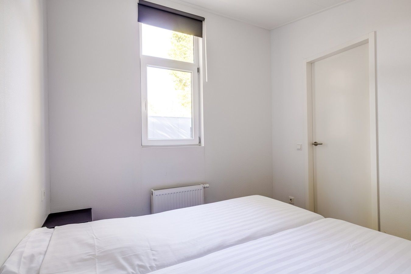 A small, single-bed room with a window, a radiator, and a door.