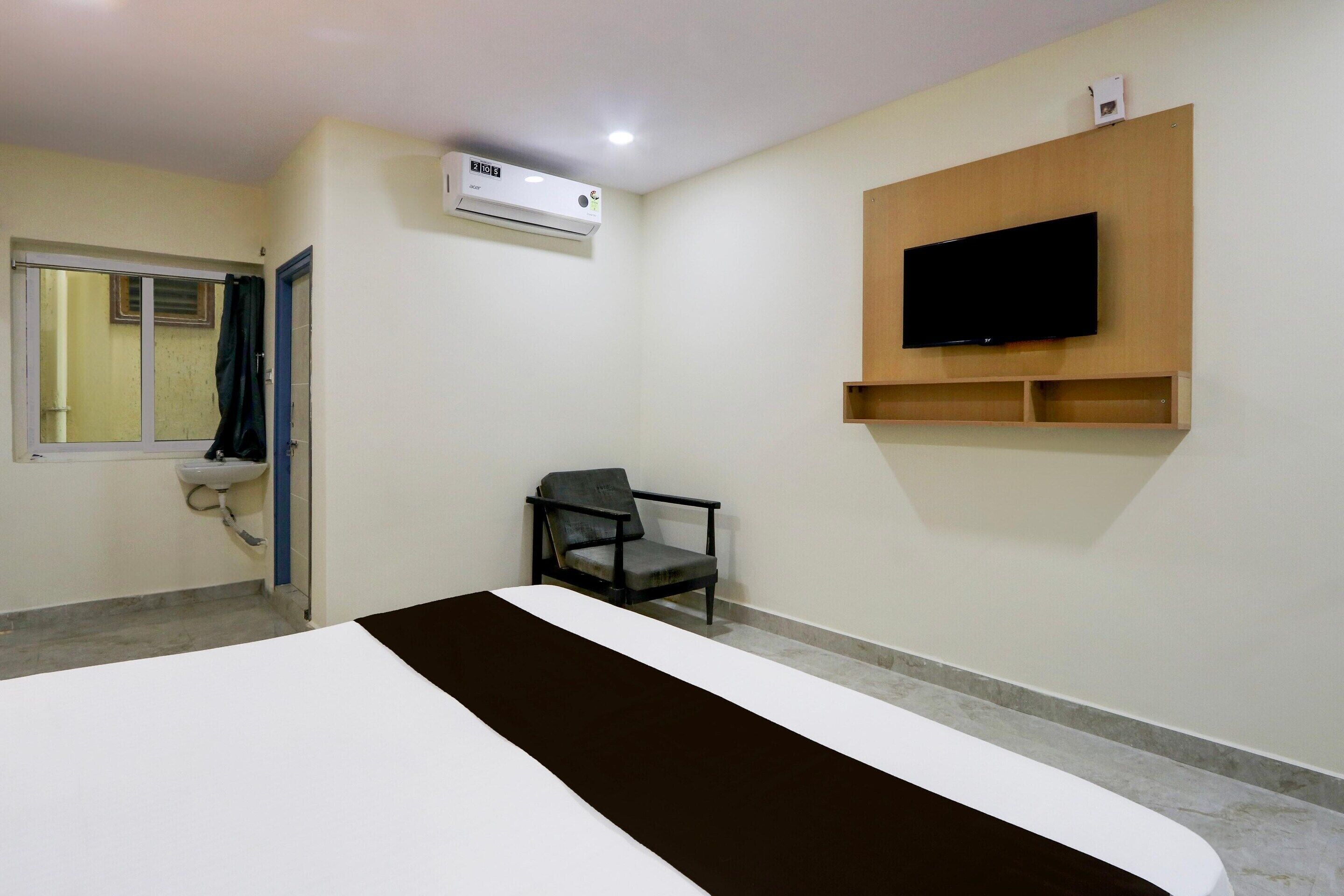 A hotel room with a bed, a chair, a TV mounted on the wall, and an air conditioning unit.