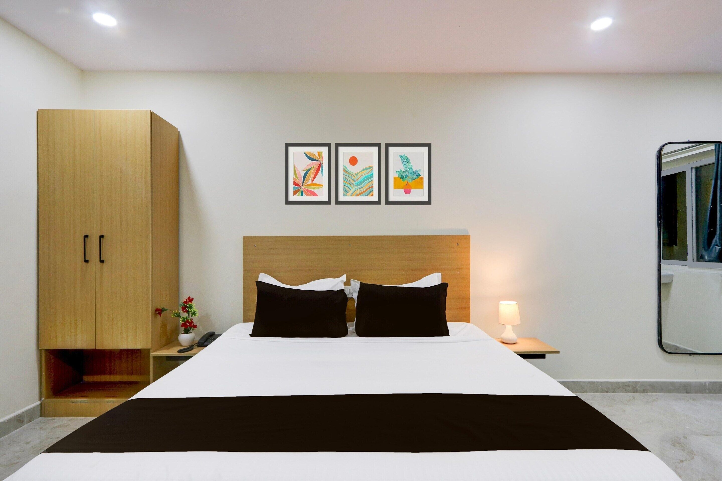 A hotel room with a bed, a wardrobe, a nightstand, a lamp, and three framed artworks on the wall.