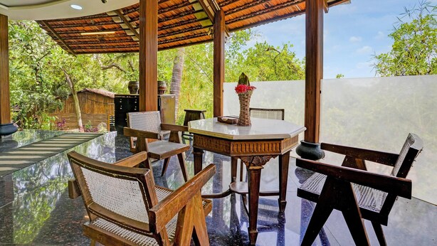 A modern outdoor dining area with a glass table, wooden chairs, and a pergola structure. - Palette Palghar Irayos Resort (Dahanu)
