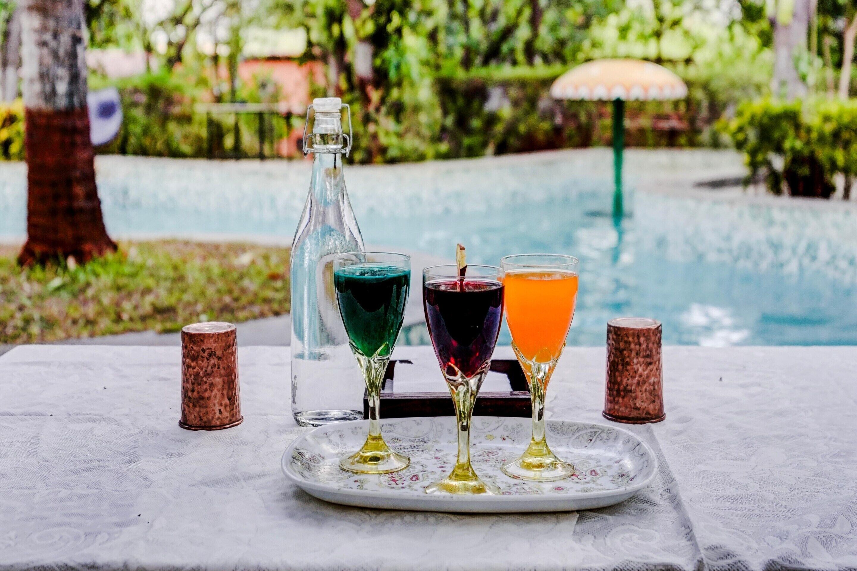 A table with a bottle and three glasses of colored drinks, a plate, and two salt shakers, set against a poolside view.