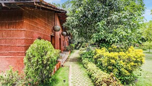A rustic wooden building with a pathway leading to it, surrounded by lush greenery and a variety of plants. - Palette Palghar Irayos Resort (Dahanu)