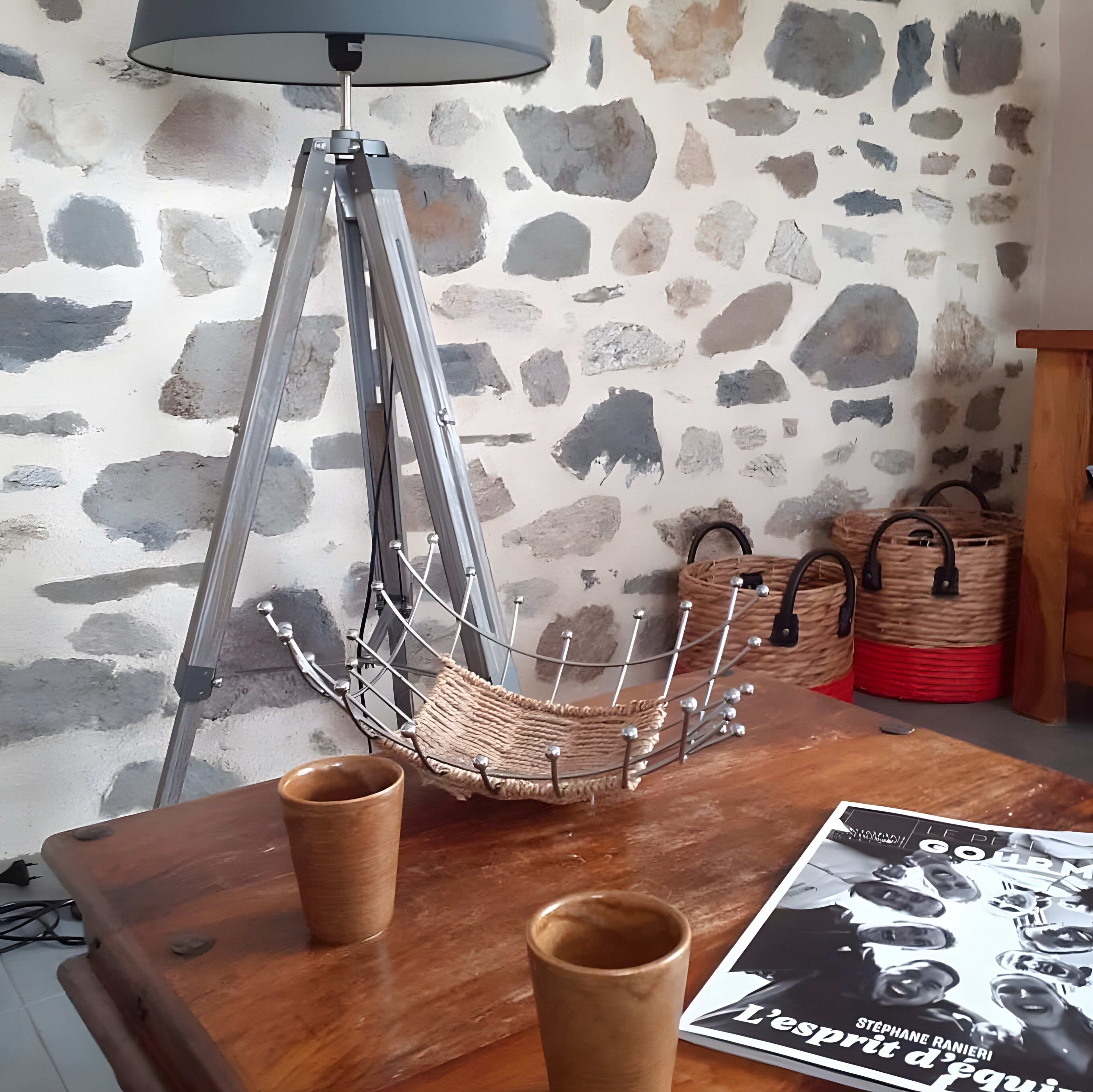 A room with a stone wall, a tripod floor lamp, a woven basket chair, two wooden cups, and a magazine on a table.