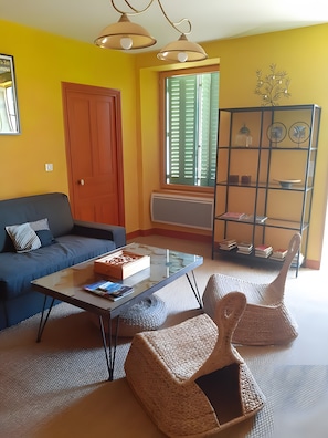 A living room with a sofa, coffee table, wicker chairs, a bookshelf, and a window with shutters. - Gîte l'oustal del pastre (Alleuze)