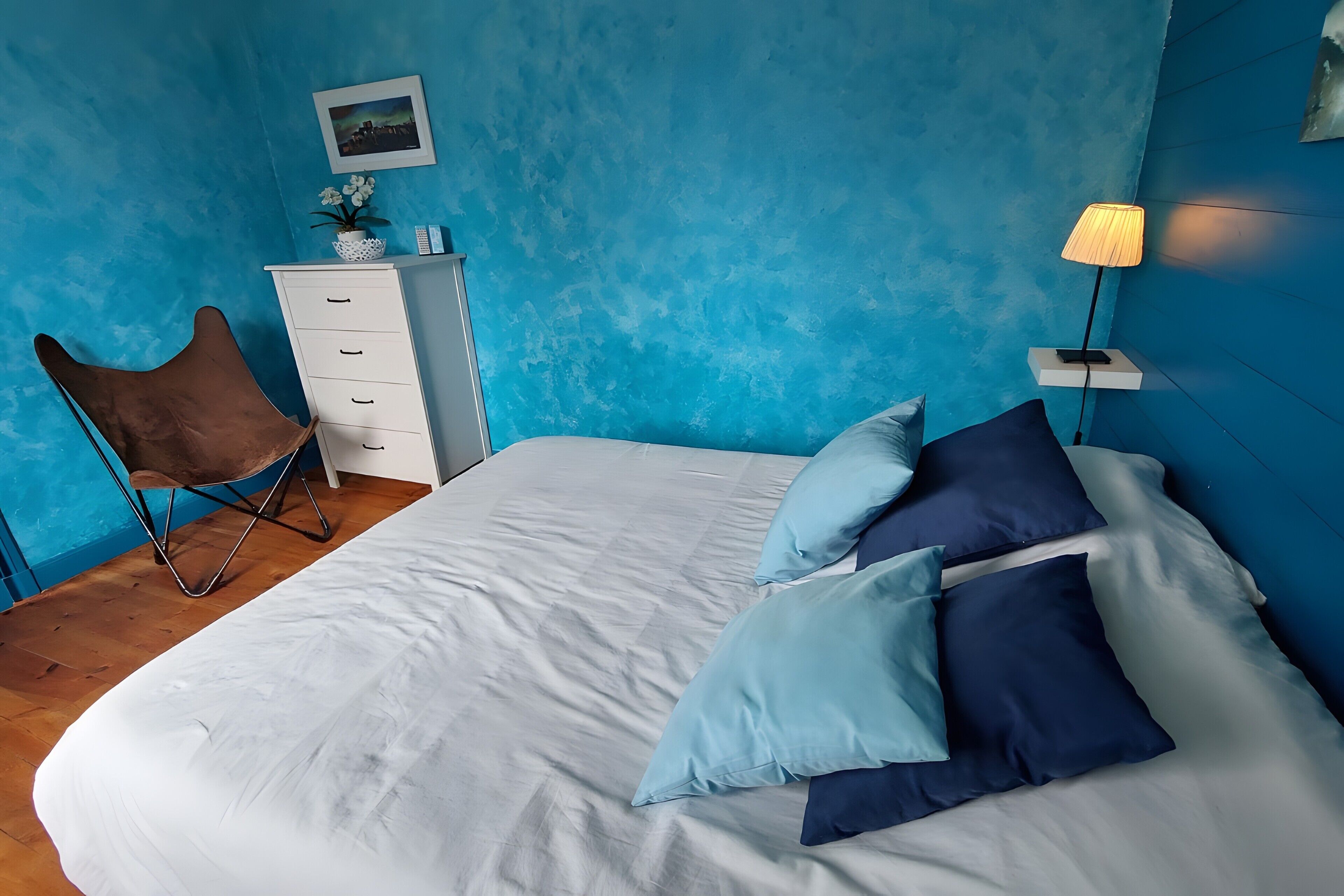 Une chambre à coucher comprenant un lit, des oreillers, une chaise, une commode, une lampe et un tableau accroché au mur.