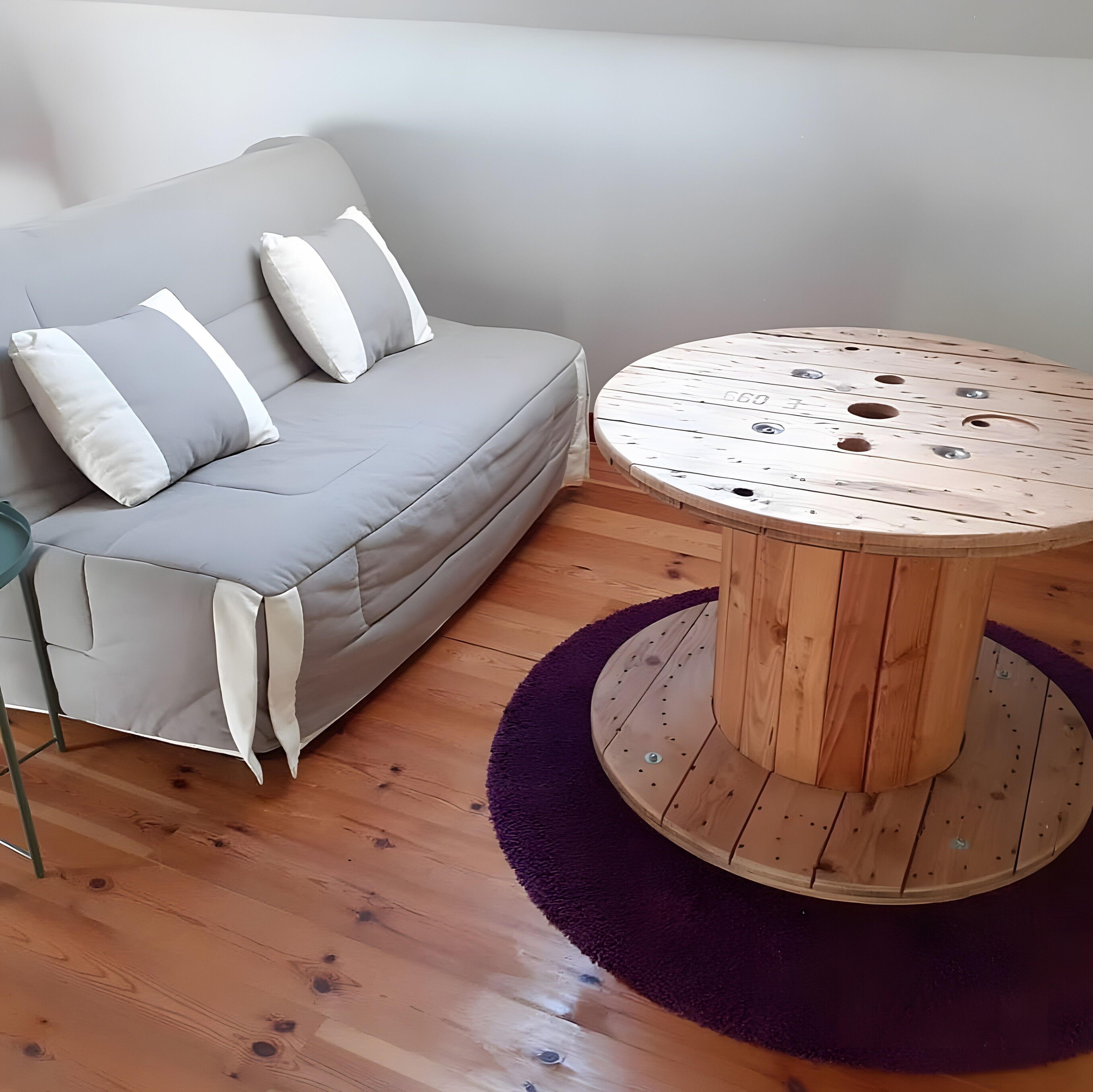 A grey sofa with white cushions, a round wooden table with holes, and a purple rug on a wooden floor.