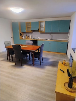 A compact kitchen with teal cabinets, a wooden dining table with four chairs, and a small countertop with a remote control and candles.