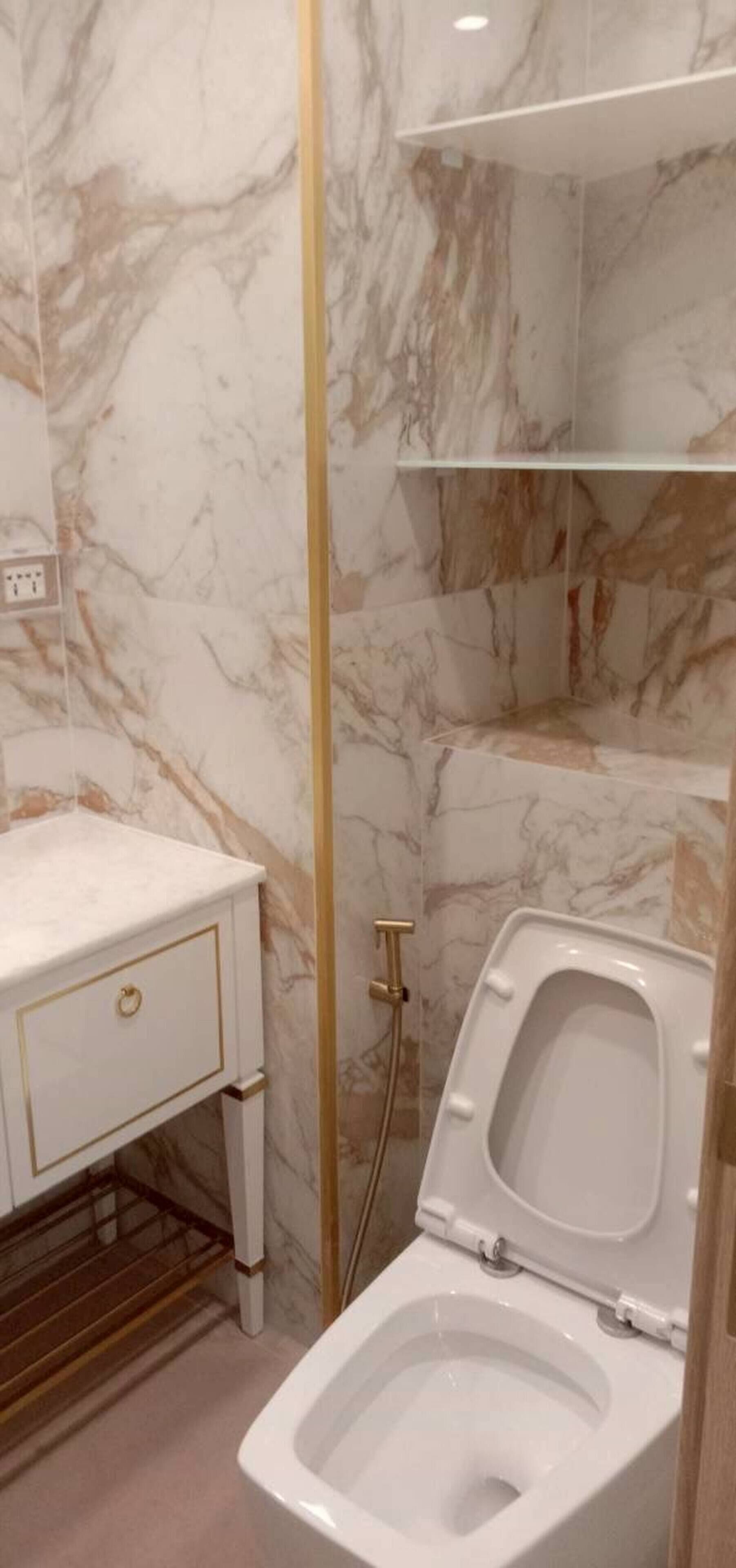 A bathroom with a marble wall, a white toilet, a white sink with gold trim, and a gold-colored shower enclosure.
