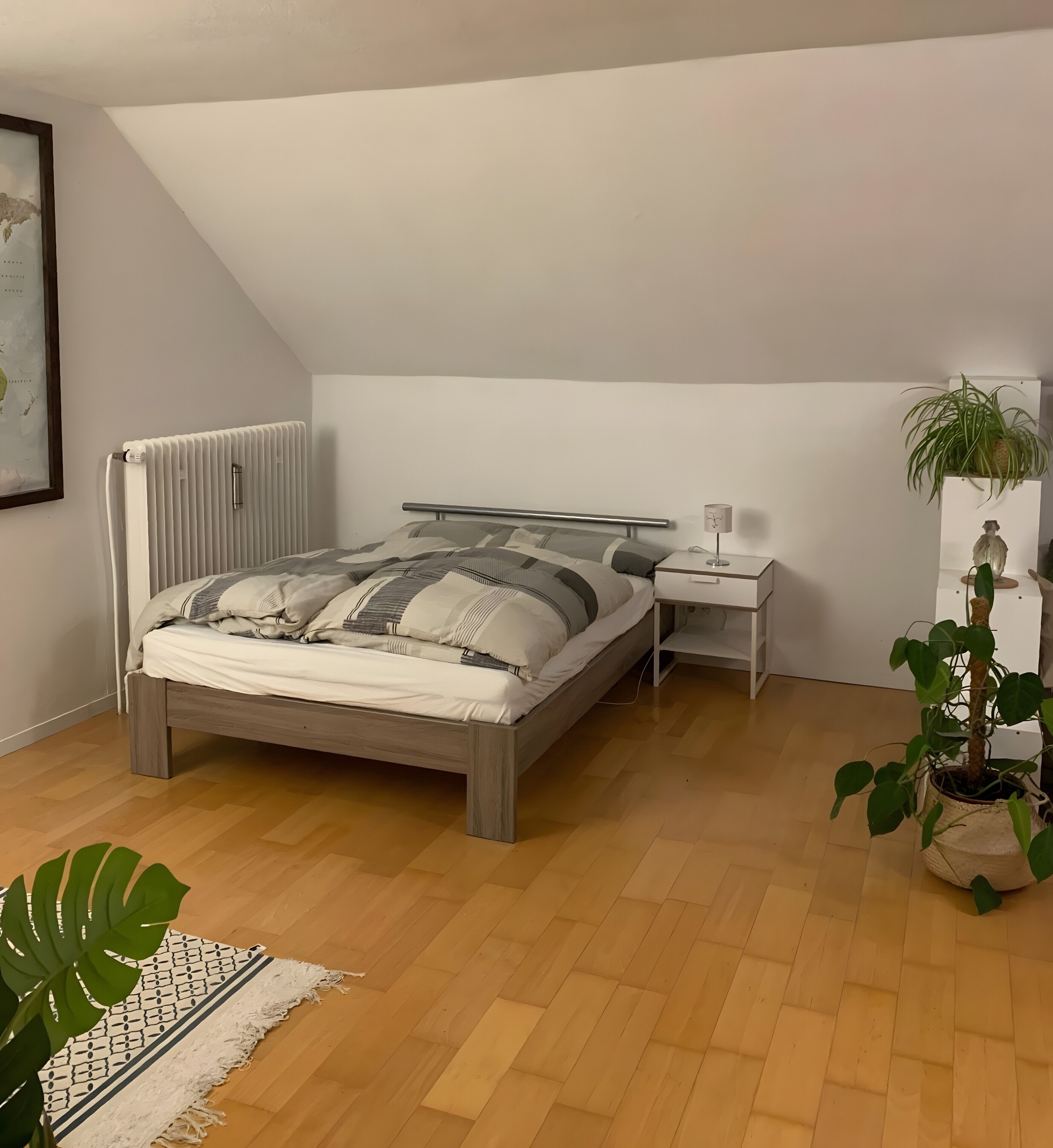 A bedroom with a bed, bedside table, lamp, radiator, potted plant, and a large window.